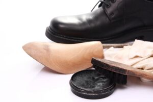 A black leather shoe is placed next to a spiral shoe tree, a shoe brush, a cloth, and an open tin of black shoe polish on a white surface.