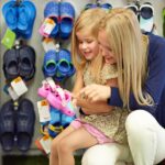 A woman and a young girl sit in a shoe store, smiling as the girl holds up a pink shoe. Behind them, a display rack showcases various colorful sandals and shoes. They're likely sharing tips to find the perfect fit among the vibrant selection.
